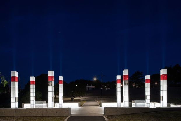 Night-time exterior view of a formal entrance or driveway framed by multiple illuminated rectangular pillars with alternating white and red horizontal bands. A paved central walkway leads through the symmetrical gateway toward a tree-lined site, with low stone or concrete walls and subdued landscape lighting.