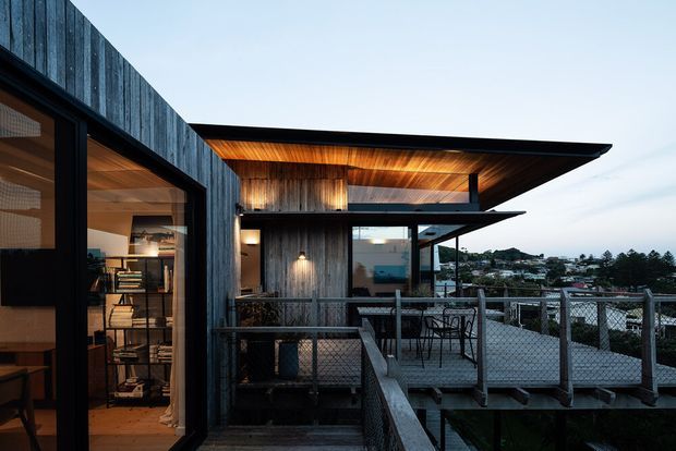 Exterior view of a contemporary, multi-level timber-clad house with dramatic projecting flat roof planes, warm timber soffits, extensive glazing, and a raised terrace with wire balustrading. Interior shelving and warm lighting are visible through the glazing, while a suburban coastal-looking landscape appears beyond.