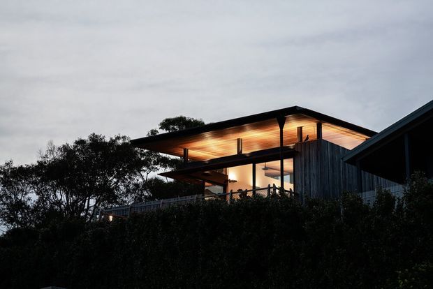 Exterior dusk view of a contemporary multi-level residence set among dense vegetation. The house features strong horizontal roof planes with deep overhangs, warm timber soffits, extensive upper-level glazing, dark vertical timber cladding, and an illuminated balcony or terrace. The silhouette and exposed roof structure create a striking, modern architectural composition against an overcast sky.