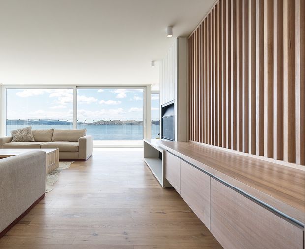 Bright contemporary living room with a calm waterfront outlook. The space features full-height glazing, a pale timber floor, a long built-in timber media console, and a vertical timber batten feature wall. Neutral upholstered sofas face the glazing and television, creating an airy, minimal interior.
