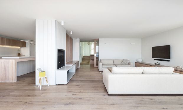Bright open-plan contemporary interior combining a kitchen, living room and central media joinery. Pale timber flooring runs throughout, with white vertical wall panelling and built-in cabinetry, light timber kitchen cabinetry, and cream upholstered sofas arranged around a wall-mounted television. A hallway and stair area extend toward the rear, while restrained furnishings and abundant neutral finishes create a clean, airy atmosphere.