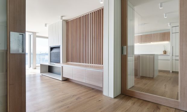 Bright contemporary open-plan interior with a pale timber floor, white walls and integrated light-wood joinery. A built-in media wall combines low cabinetry, a television and a vertical timber slat feature panel. The adjoining kitchen is visible through a large timber-framed glazed opening, with a central island, pale cabinetry and simple recessed or surface-mounted lighting. The room has a clean, airy and refined appearance, with a glimpse of expansive glazing and an outdoor view in the background.
