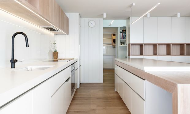 Bright contemporary kitchen with a long white island and matching perimeter cabinetry, pale timber accents, vertical wall panelling, integrated shelving, and linear ceiling lighting. The space has warm timber flooring and a clean, streamlined layout with a secondary pantry or utility area visible at the rear.