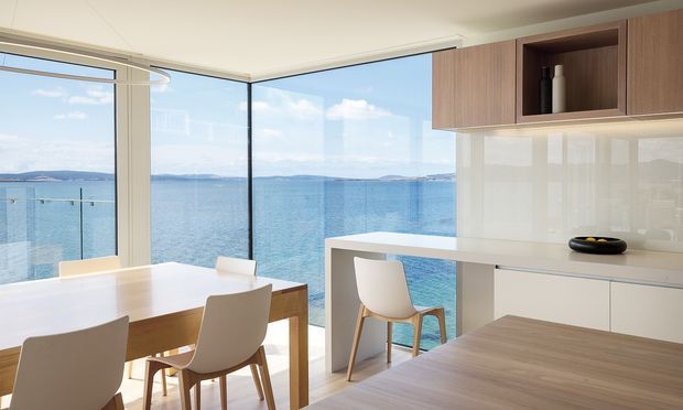 Bright contemporary kitchen and dining area with floor-to-ceiling corner glazing overlooking blue water and distant coastline. The room features a light timber dining table, white molded chairs, a white island or breakfast bar, glossy white splashback, white lower cabinetry, and warm timber overhead joinery. A glass balustrade is visible outside the glazing, creating an airy waterfront setting.
