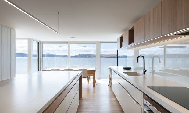 Bright contemporary kitchen with a large central island and a parallel run of white cabinetry and benchtop. Warm timber overhead cabinetry and light timber flooring add natural contrast. Full-height glazing opens the kitchen and dining area to expansive waterfront views, with minimalist linear lighting and streamlined black and metallic fixtures.
