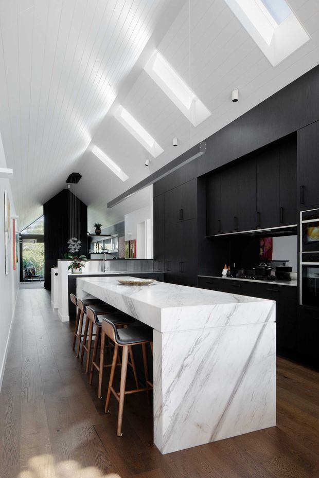 A bright, elongated contemporary kitchen with a large white marble island and integrated seating for four. The kitchen features extensive matte black full-height cabinetry, dark timber flooring, integrated appliances, and a white paneled pitched ceiling with multiple skylights. A glazed area and living space are visible beyond the kitchen, while pendant and recessed lighting add illumination.
