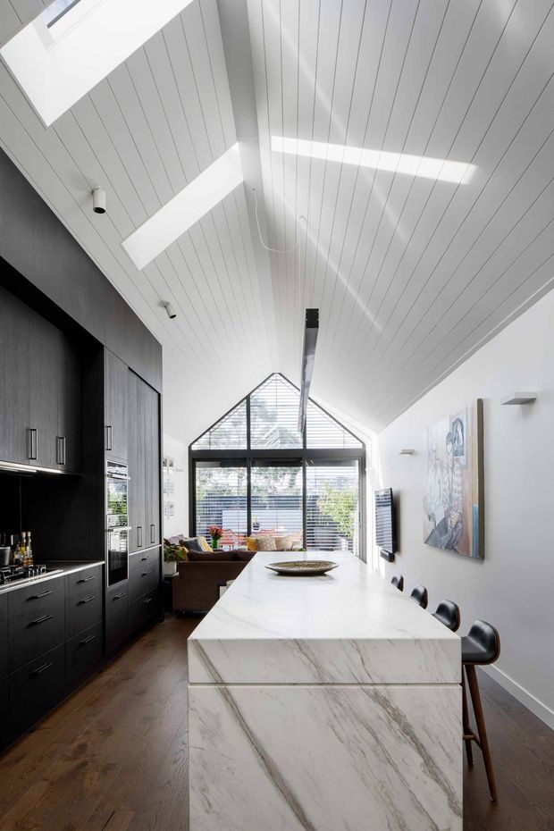 A bright, elongated open-plan kitchen and living space beneath a dramatic pitched, white-painted timber-panelled ceiling. The kitchen features dark charcoal cabinetry, integrated appliances, a large white marble island with waterfall ends, and black counter stools. At the far end, full-height glazing with horizontal blinds and a triangular gable window brings in abundant natural light. Warm timber flooring, contemporary artwork, and a compact living area complete the interior.