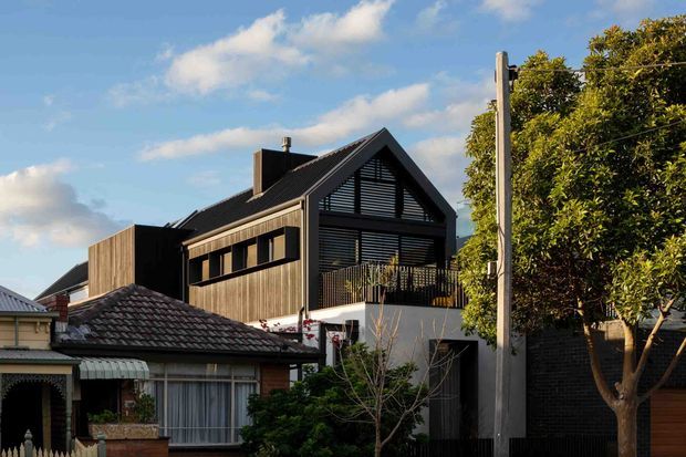 Exterior view of a contemporary multi-level residence with a steep gable roof, dark vertical cladding, extensive horizontal louvres, and a projecting upper balcony. The home is nestled among mature suburban trees and sits beside older neighbouring homes, creating a contrast between new and established architecture.