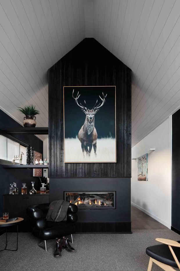A dramatic, moody living room centered on a black fireplace wall with vertical dark timber paneling and a large deer artwork above the fire. The room has a steep white timber-lined vaulted ceiling, built-in black shelving and bar storage, a black leather lounge chair, dark flooring, and a carpeted seating area. A hallway extends to the right.