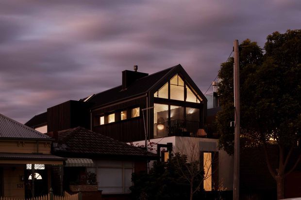 Twilight exterior view of a contemporary multi-level house in an urban residential setting. The dark-clad dwelling has a prominent steep gabled roof, large illuminated upper-level glazing with horizontal slatted screening, and a mix of modern and neighbouring traditional homes in the foreground.