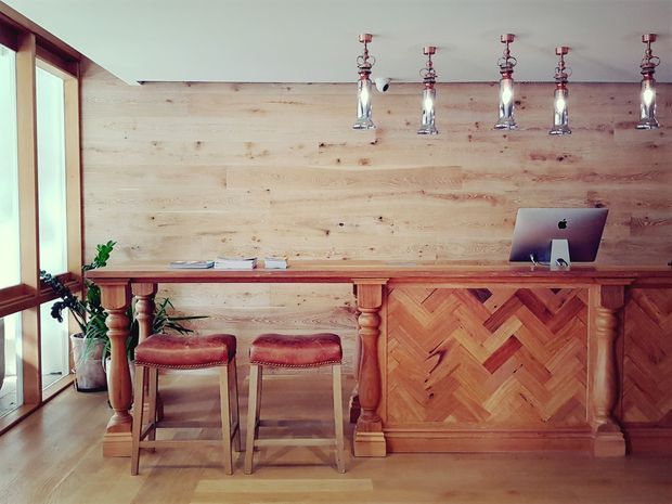 A warm, rustic reception or service counter area featuring a timber-clad wall, substantial timber counter with turned columns, herringbone timber paneling, two leather-topped stools, vintage-style pendant lights, and a desktop computer. Large timber-framed glazing is visible on the left, with indoor plants adding greenery.