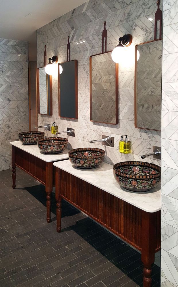A richly detailed bathroom with a long run of individual washbasins set on dark timber vanity consoles. The room features patterned marble wall tiles in a chevron arrangement, white stone benchtops, dark floor tiles, ornate decorative vessel basins, copper-toned framed mirrors, and black-and-brass wall sconces. The repeated vanity layout suggests a commercial or hospitality bathroom.