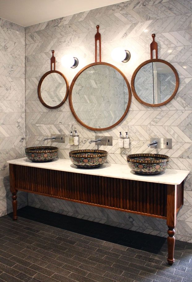 A richly detailed bathroom vanity with three ornate patterned vessel basins set on a long dark timber console with a pale stone benchtop. The walls are clad in light grey marble-look chevron tiles, complemented by three round timber-framed mirrors with decorative vertical supports and contemporary wall sconces. Dark textured floor tiles and vintage-inspired fixtures create an eclectic, luxurious character.