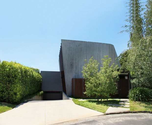 Street-facing view of a contemporary residential house with a sculptural curved concrete facade, dark recessed garage, vertical timber screening, and a broad pale concrete driveway. Dense hedges and established trees partially frame the minimalist building.