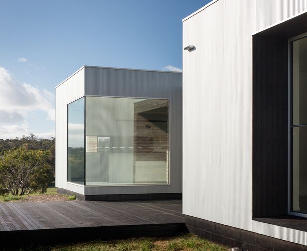 Exterior architectural photograph of a contemporary, flat-roofed building composed of crisp white corrugated cladding and dark recessed window and door openings. A glazed projecting volume with floor-to-ceiling windows sits on a dark timber deck, with bushland visible beyond. The composition emphasises clean geometric forms, strong horizontal and vertical lines, and a restrained monochrome material palette.