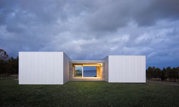 A low, modern single-storey building sits in an open grassy landscape under a dramatic cloudy sky. Two large white rectangular volumes flank a recessed central entry or glazed opening, framed with warm timber cladding and illuminated from within. The minimalist composition has crisp geometric forms and a strong horizontal layout.