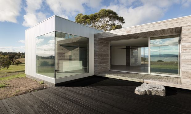 Exterior view of a low-slung contemporary residence on a rural or semi-rural site. The building combines a crisp white rectangular volume with extensive floor-to-ceiling glazing and a weathered horizontal timber-clad wing. A broad dark timber deck forms the foreground terrace, while the open-plan interior is visible through the glazing. The composition emphasizes strong rectilinear geometry, indoor-outdoor connection, and integration with the surrounding landscape.