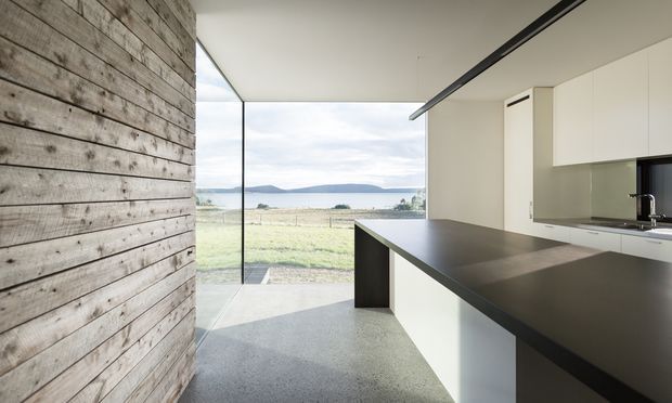 A minimalist contemporary kitchen with a long dark island and white cabinetry, opening through full-height glazing to a rural waterfront landscape. The space features a prominent weathered timber feature wall, polished light concrete flooring, a restrained white and charcoal palette, and a slim linear ceiling light.
