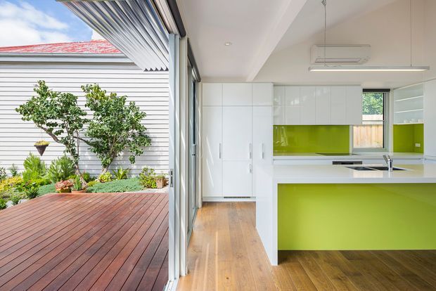 Bright contemporary kitchen opening directly onto a timber deck and landscaped courtyard through wide glazed folding or sliding doors. The kitchen features glossy white full-height cabinetry, a vivid lime-green splashback and island front, timber flooring, stainless-steel appliances and a linear pendant light. The adjoining exterior includes horizontal weatherboard cladding, a red roof edge, timber decking and planted garden beds.