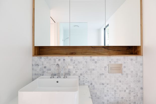 A bright, compact bathroom vanity featuring a white rectangular basin with wall-mounted chrome tapware, a mirrored cabinet framed by a thick natural timber surround, and a pale grey-white mosaic tiled splashback. The clean-lined design combines warm timber with crisp white and cool neutral finishes.