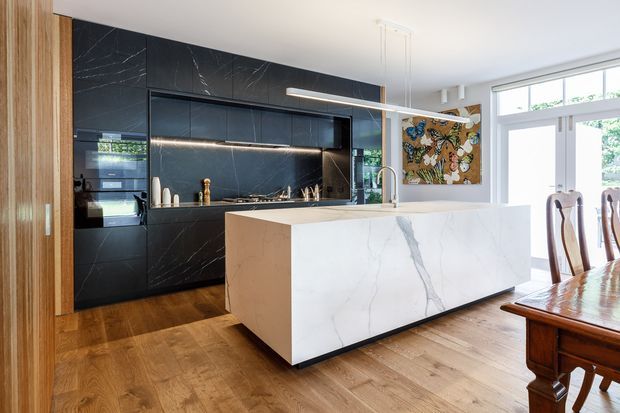 Bright contemporary kitchen with a large white marble-look island, dark charcoal veined cabinetry and splashback, integrated appliances, timber flooring, and a linear pendant light. A traditional timber dining table and chairs sit beside the kitchen, with large windows bringing in natural light.