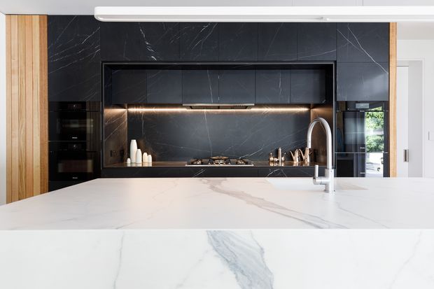 Contemporary kitchen featuring a large white stone island with subtle grey veining, a dark marble-look splashback and integrated cooking zone, black appliances and cabinetry, warm timber wall panels, and under-cabinet lighting. The composition is clean-lined and high contrast, with a generous island and concealed storage.