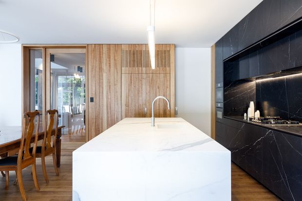 Contemporary kitchen with a large white stone island and integrated sink, dark marble-effect cabinetry and splashback, warm timber flooring, and a timber-panelled wall with concealed storage. A dining area and glazed doors are visible to the left. The space is bright, refined, and strongly contrasted between light stone, warm timber, and dark cabinetry.
