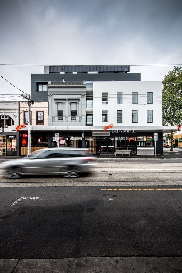 Street-facing mixed-use urban building with a contemporary multi-storey addition above retained ornate historic shopfront architecture. The facade combines dark charcoal cladding, crisp white rendered walls, tall narrow windows, and a ground-floor burger restaurant frontage. Viewed across a busy road with a motion-blurred car, overhead utility wires, and neighboring commercial buildings.