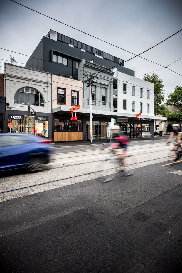 Street-level view of a dense urban commercial streetscape with attached multi-storey shopfront buildings. The frontage combines older masonry and ornate heritage-style facade elements with newer dark and white upper-level additions. Ground-floor businesses include a gym, retail storefronts, and a healthy burger restaurant. Tram or trolley wires, street tracks, moving vehicles, and cyclists emphasize the active urban setting.