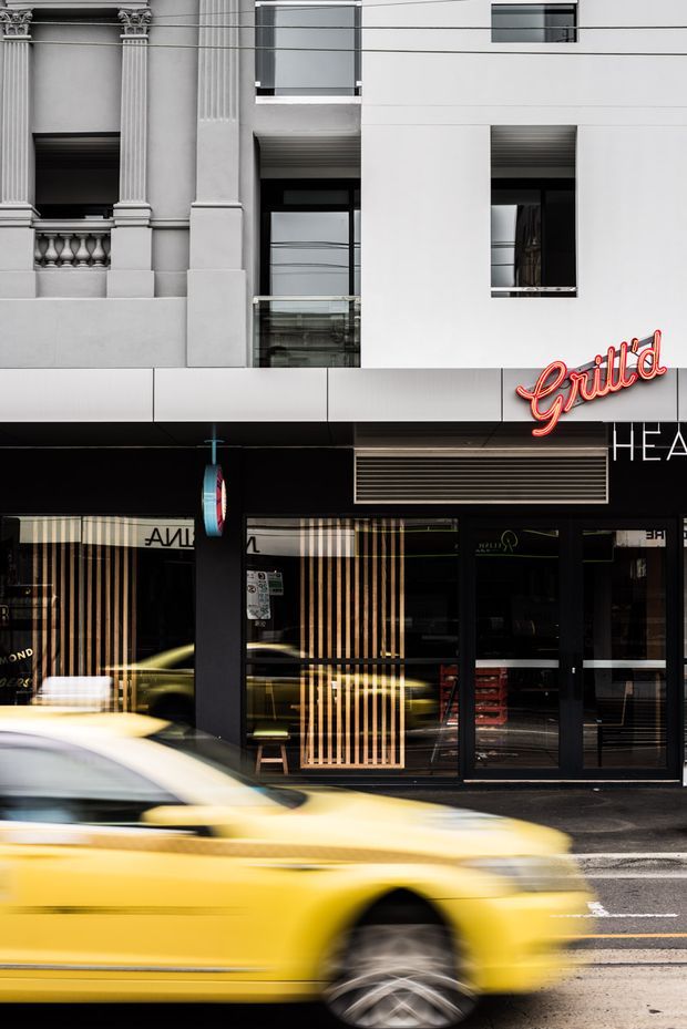 Street-level view of a contemporary urban commercial frontage integrated with an older ornate heritage facade. The ground floor has dark glazing, vertical timber-look screening, black framing, a projecting illuminated red “Grill'd” sign, and a louvred ventilation panel. A blurred yellow taxi passes in the foreground.