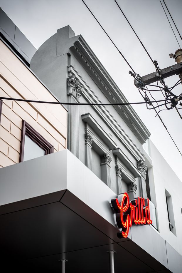 Upward-looking street view of a commercial building facade combining a highly ornate classical-style frontage with contemporary white cladding and a projecting dark canopy. A bright red neon restaurant sign is mounted above the entrance, with overhead utility wires and a pole crossing the overcast sky.