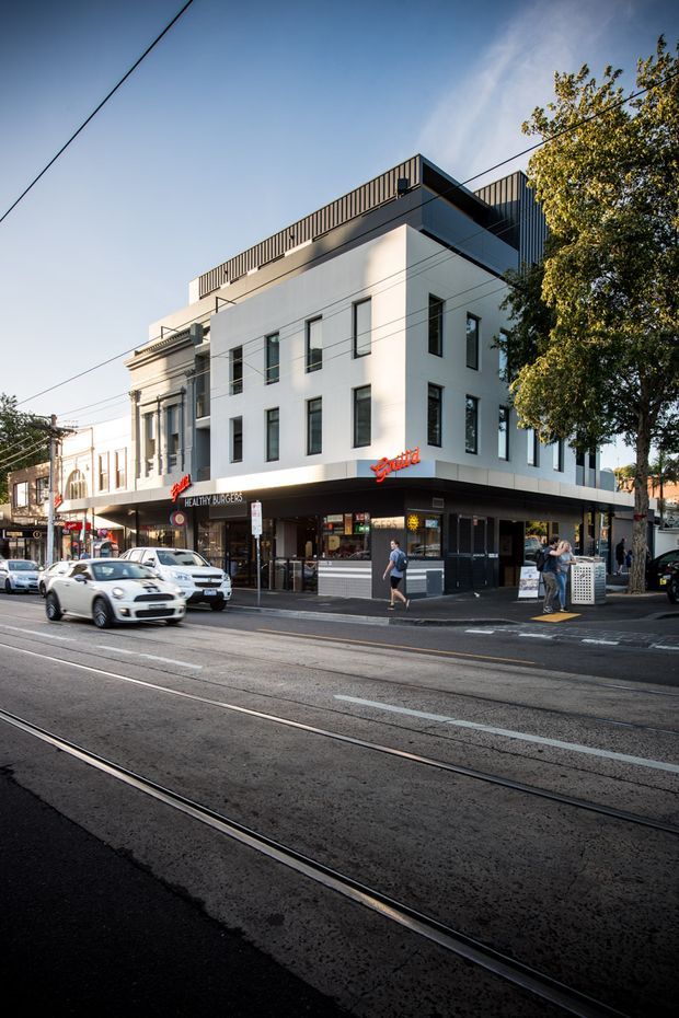 Street-level view of a contemporary multi-storey mixed-use urban building with a white rendered facade, narrow vertical windows, rooftop terrace railings, and ground-floor retail and food outlets. The building occupies a prominent corner on a tram-lined commercial street, with adjacent older masonry buildings and mature street trees.
