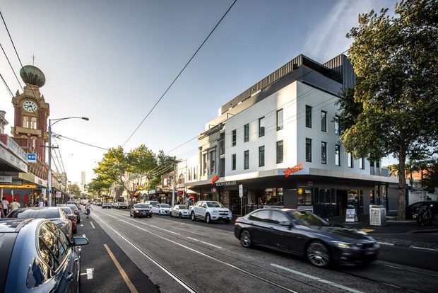 Street-level urban view of a contemporary multi-storey commercial building with a white rendered facade, dark ground-floor shopfronts, narrow vertical windows, and rooftop terraces. The building sits within an active streetscape with tram tracks, overhead wires, retail signage, parked cars, and adjacent older commercial architecture.
