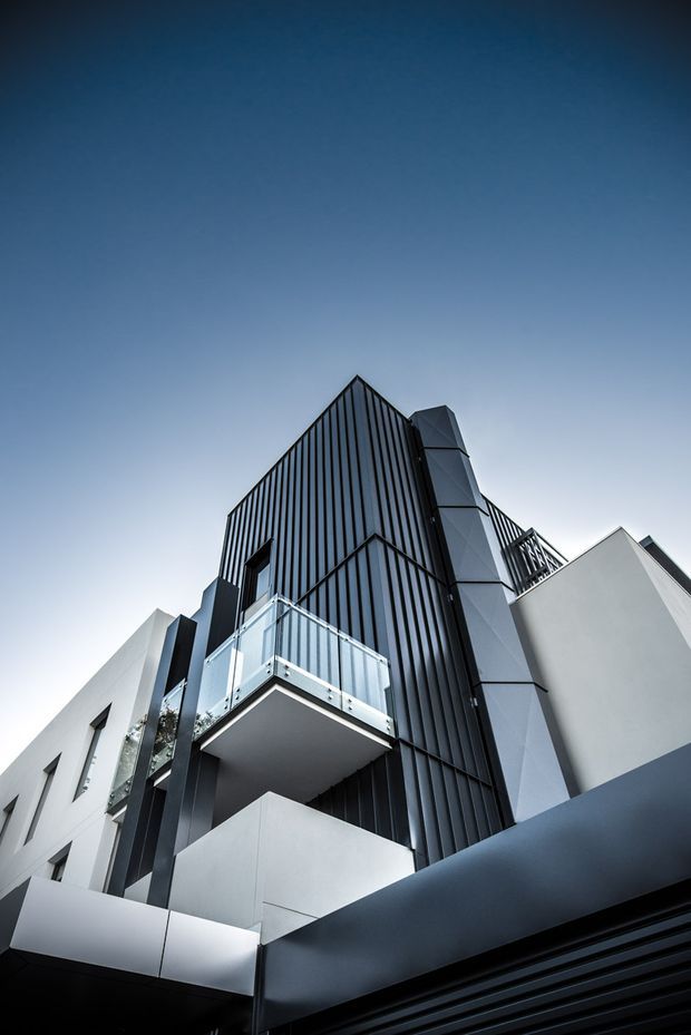 Low-angle exterior photograph of a contemporary multi-storey residential building. The facade combines dark vertically ribbed cladding with crisp white rendered volumes, projecting balconies with clear glass balustrades, and angular geometric forms. The image is dominated by the building envelope against a clear blue sky.