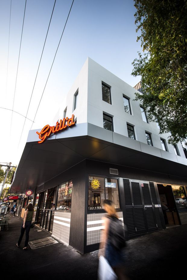 Street-level view of a contemporary multi-storey commercial building on a busy urban corner. The lower level has a dark charcoal facade, black soffit and louvred screening, glazed shopfronts and a prominent illuminated red-orange “Grill’d” sign, while the upper levels are crisp white rendered volumes with simple rectangular windows. Pedestrians, street infrastructure and mature trees frame the building.