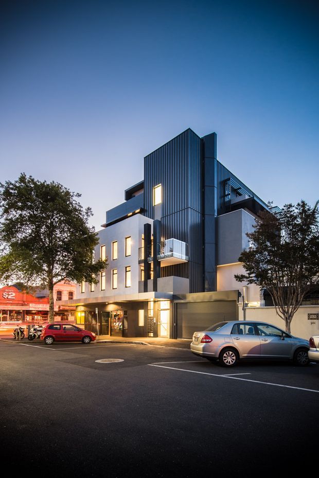 A dusk street-level view of a contemporary multi-storey urban building with a sharply articulated dark vertical metal-clad facade, pale rendered walls, projecting balconies, illuminated windows, ground-floor glazing, and an integrated roller-door garage. The building sits directly along a developed city street with adjacent commercial buildings and parked cars.