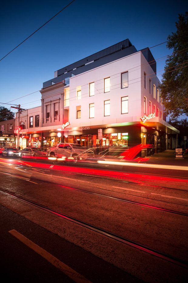 Twilight street-level exterior photograph of a multi-storey urban commercial building occupied by a brightly illuminated Grill'd burger restaurant. The modern pale facade incorporates tall rectangular windows, a projecting dark ground-floor canopy, rooftop metal screening, and retained ornate detailing on the adjoining older building. Tram tracks, overhead wires, street lighting, and blurred vehicle light trails create a dynamic city streetscape.