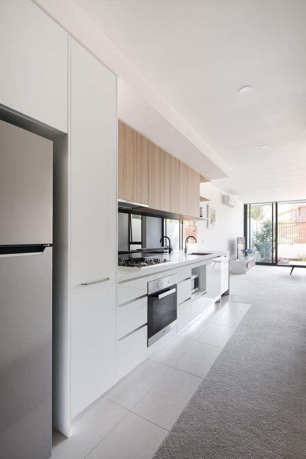 Bright, minimalist open-plan kitchen viewed along a long wall of white cabinetry. The kitchen features integrated stainless-steel appliances, a white benchtop, pale timber overhead cupboards, black-framed window and splashback details, and a light tiled floor transitioning to a grey carpeted living area. Full-height glazing at the rear opens toward an outdoor space.