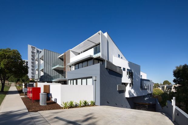 Exterior view of a contemporary multi-level residential building on a sloping urban site. The building has angular white and grey volumes, corrugated or ribbed metal cladding, projecting balconies, extensive dark-framed glazing, and a partially concealed lower-level garage. A concrete driveway and pedestrian path frame the foreground under a clear blue sky.
