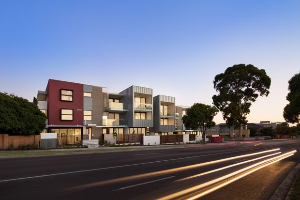 Street-level dusk view of a contemporary multi-storey apartment development with varied rectangular volumes, dark grey vertical cladding, muted red facade panels, balconies, large windows, and ground-level entries. The building fronts a broad urban roadway with vehicle light trails, mature trees, and low-rise surroundings.