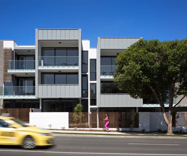 Street-facing view of a contemporary multi-storey residential apartment building with flat rooflines, projecting box forms, grey metal-look facade panels, extensive dark glazing, glass balconies, and vertical timber-look screening. A landscaped frontage and street are visible, with a blurred vehicle in the foreground.