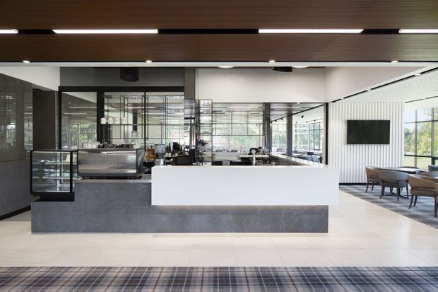 A bright contemporary commercial hospitality interior, likely a café, restaurant, or hotel lobby bar. The foreground is dominated by a large white service counter with dark grey stone-faced cabinetry and a glass display case. Behind it are coffee and bar-service facilities with black-framed glazed partitions. The space has a dark timber slatted ceiling with recessed linear lighting, white tiled flooring, a vertically ribbed white feature wall, a wall-mounted screen, and a small seating area by expansive windows.