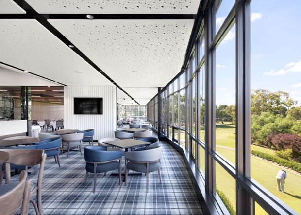 A bright contemporary hospitality lounge or dining space with multiple upholstered tables and chairs arranged along a full-height glazed facade. The room features a patterned carpet, white linear wall panelling, a black-framed suspended acoustic ceiling with recessed lighting, and views over landscaped grounds through large windows.