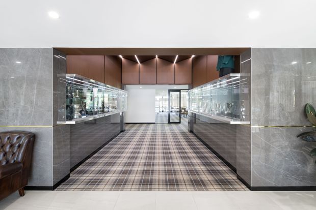 A sophisticated commercial interior corridor or lobby with symmetrical glass display cases on both sides, dark grey marble-look wall panels, warm wood-toned overhead cabinetry or wall cladding, and a patterned plaid carpet runner. Recessed linear lighting creates a refined, gallery-like atmosphere, with a glazed doorway visible at the far end and partial leather seating and planting at the edges.