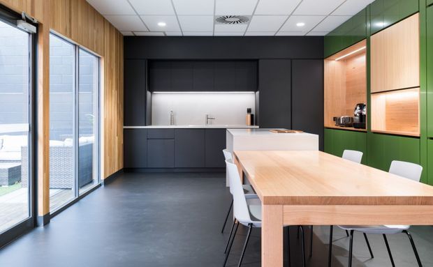 A contemporary open-plan kitchen and dining area with dark matte cabinetry, a white island, and a large light timber dining table. Full-height glazed doors line one side, while a green built-in wall with warm timber shelving and integrated appliances adds contrast. The space has a clean suspended acoustic ceiling and dark flooring.