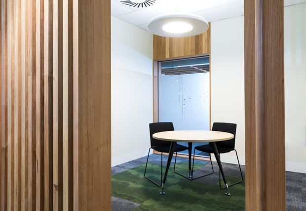 A compact contemporary meeting room viewed through a timber-framed opening. The room features vertical timber wall battens, warm timber joinery around a glazed opening, a round light-wood table, black chairs, a patterned green and grey carpet, white walls and ceiling, and a suspended circular pendant light. The space appears to be a small commercial office meeting room.