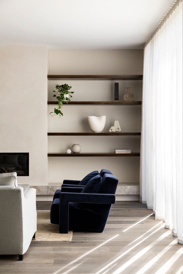 A refined contemporary living room with a built-in recessed shelving wall, dark timber shelves, neutral textured wall finish, light timber flooring, and a stone-clad low ledge. A deep navy upholstered armchair faces a partially visible light sofa and wall-mounted fireplace. Full-height sheer curtains filter strong daylight from the right, creating bright linear shadows across the floor. Minimal decorative objects and a trailing plant add understated warmth.