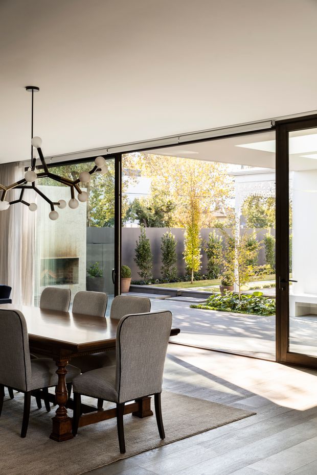 A refined dining room with a large timber dining table and upholstered dining chairs, opening through expansive dark-framed glazed doors to a landscaped courtyard. The room features pale timber flooring, a neutral rug, sheer curtains, a marble or stone fireplace surround, and a sculptural branching pendant light. Strong indoor-outdoor connection and abundant natural light create a contemporary, elegant atmosphere.