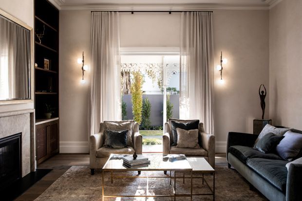 Elegant, warmly lit living room with a restrained classic interior. The room features a marble-trimmed fireplace, dark built-in shelving, tall curtains framing a glazed garden-facing opening, two upholstered armchairs, a dark sofa, a glass-topped brass coffee table, and a neutral textured rug. Traditional architectural detailing is paired with refined contemporary furnishings.