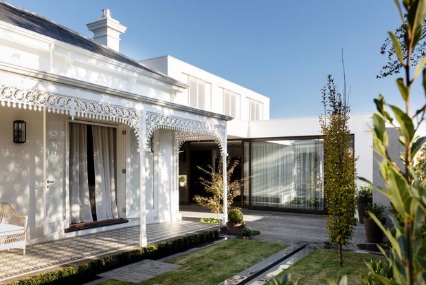 A bright residential courtyard showing a heritage-style white facade with ornate cast-iron lacework, decorative verandah detailing, tall windows and a chimney, connected to a crisp contemporary white extension with large glazed walls. The landscaped courtyard includes formal paving, narrow garden beds, clipped shrubs and young trees.